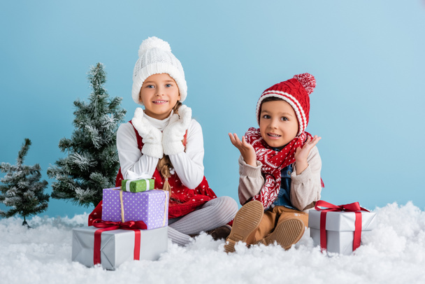 children in winter outfit sitting on snow and gesturing near presents and fir isolated on blue - Fotografie, Obrázek