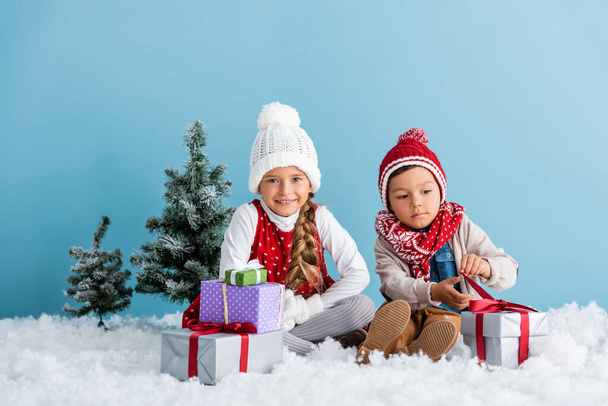 niños en traje de invierno sentados en la nieve cerca de regalos y abetos aislados en azul - Foto, Imagen