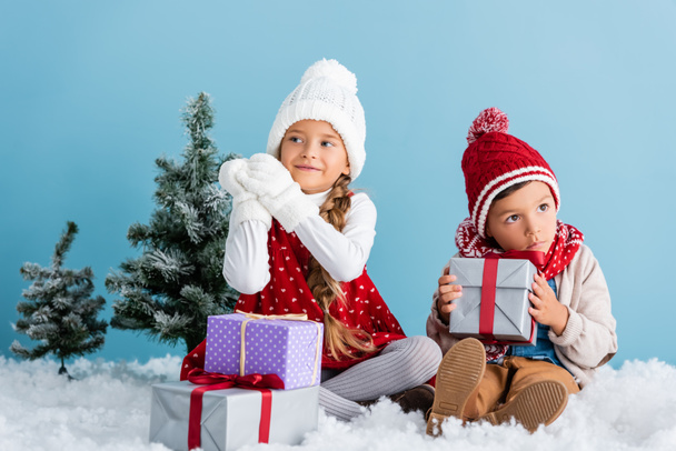 children in winter outfit sitting on snow near firs and presents isolated on blue - Fotografie, Obrázek