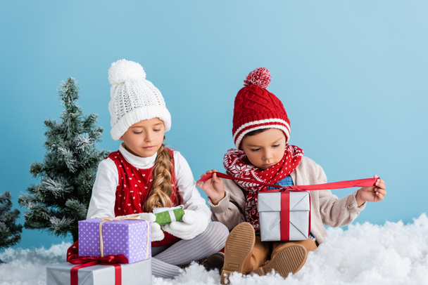 Kinder im Winteroutfit sitzen auf Schnee in der Nähe von Tannen und halten Geschenke isoliert auf blauem Grund. - Foto, Bild