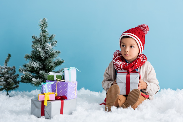 garçon en chapeau et tenue d'hiver assis sur la neige près des cadeaux et des arbres de Noël isolé sur bleu - Photo, image