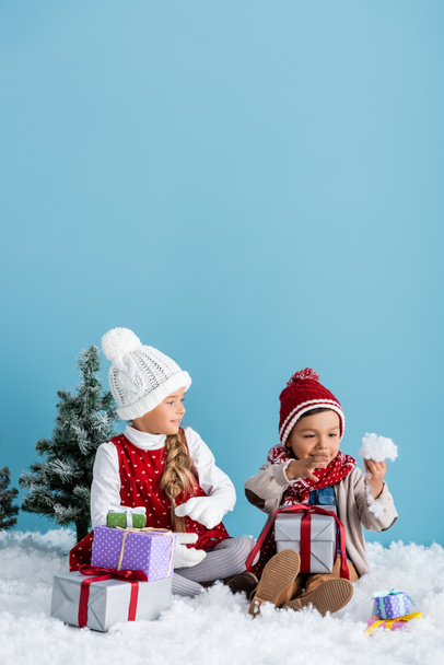 παιδί με χειμωνιάτικη στολή κάθεται κοντά σε δώρα και κοιτάζει τον αδελφό του κρατώντας χιόνι απομονωμένο στο μπλε - Φωτογραφία, εικόνα