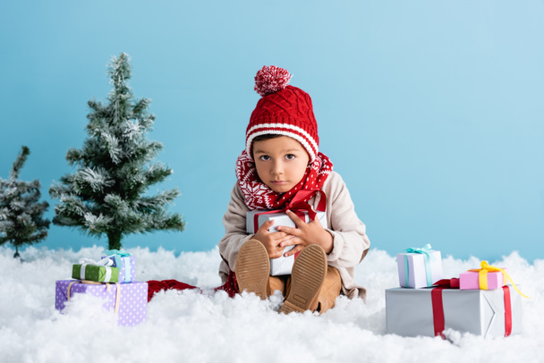 boy in hat and winter outfit sitting on snow and holding present near christmas trees isolated on blue - Photo, Image