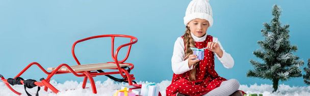 panoramic shot of kid in hat and winter outfit sitting on snow and holding present near sleigh isolated on blue - Photo, Image