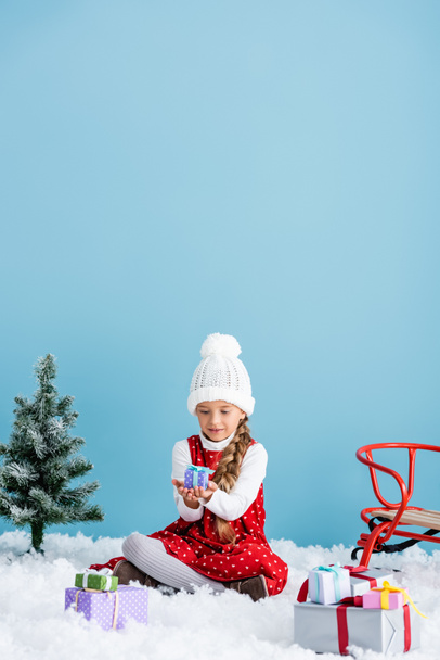 kid in hat and winter outfit sitting on snow and holding present near sleigh isolated on blue - Photo, Image
