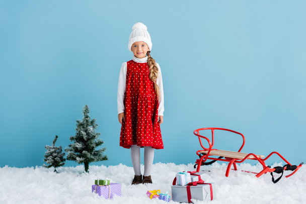 girl in hat and winter outfit standing on snow near presents and sleigh isolated on blue - Photo, Image