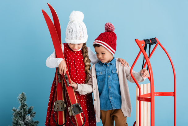 κορίτσι στο χειμώνα στολή κρατώντας σκι κοντά στον αδελφό με μαγεία στο μπλε  - Φωτογραφία, εικόνα