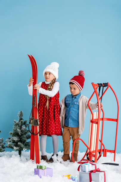 κορίτσι σε χειμερινή στολή κρατώντας σκι κοντά αγόρι στέκεται κοντά σε μαγεία και παρουσιάζει στο χιόνι απομονώνονται σε μπλε  - Φωτογραφία, εικόνα