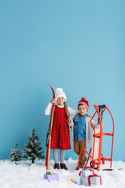 child in winter outfit holding skis and hugging boy standing near sleight isolated on blue  - Photo, Image