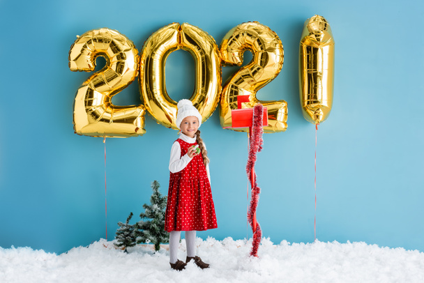kid in winter outfit holding present near mailbox and balloons with numbers while standing on snow on blue  - Photo, Image