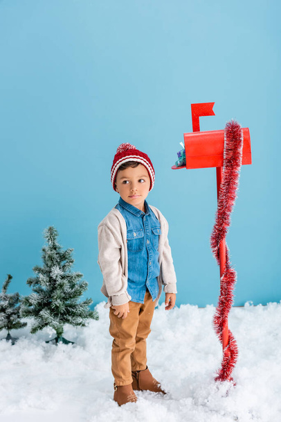 мальчик в шляпе и зимнем наряде стоит рядом с красным почтовым ящиком с подарком на синем  - Фото, изображение