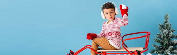 horizontal image of boy in winter earmuffs and sweater sitting in sleigh and holding snowball isolated on blue - Foto, imagen