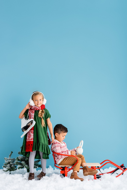 дитина в зимових вухах і шарфі стоїть з льодовими ковзанами біля хлопчика на санях на синьому
 - Фото, зображення
