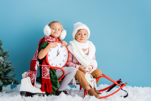 дети в зимнем наряде и шарфах сидят на санях рядом с часами, сосновые и ледяные коньки на голубом - Фото, изображение