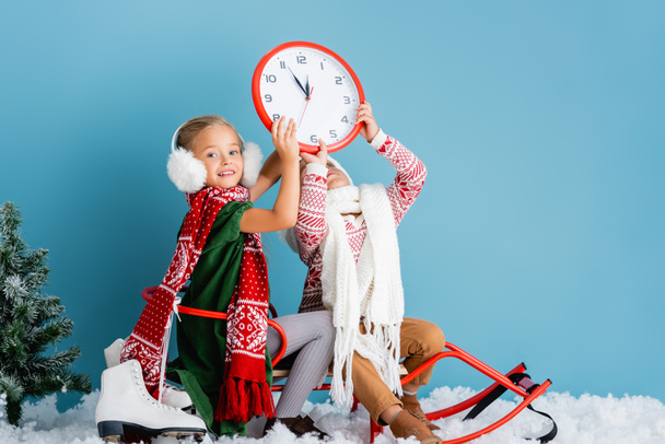dzieci w stroju zimowym siedzi na saniach i trzymając zegar w pobliżu sosny i łyżwy na niebiesko - Zdjęcie, obraz