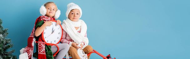 poziome uprawy dzieci w stroju zimowym i szaliki siedzi na saniach z zegarem w pobliżu sosny na niebiesko - Zdjęcie, obraz