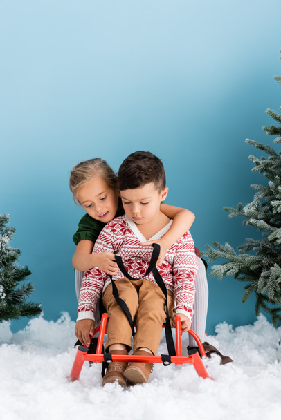 children sitting in sleigh on white snow near green pines on blue - Foto, afbeelding