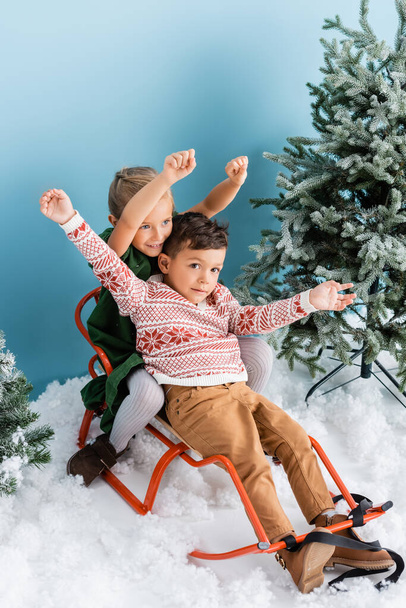 bambini eccitati con le mani sopra le teste seduti in slitta sulla neve bianca vicino a pini verdi su blu - Foto, immagini
