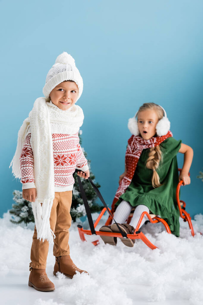 selective focus of boy in scarf and hat giving a ride to sister on sleigh on blue - Foto, imagen