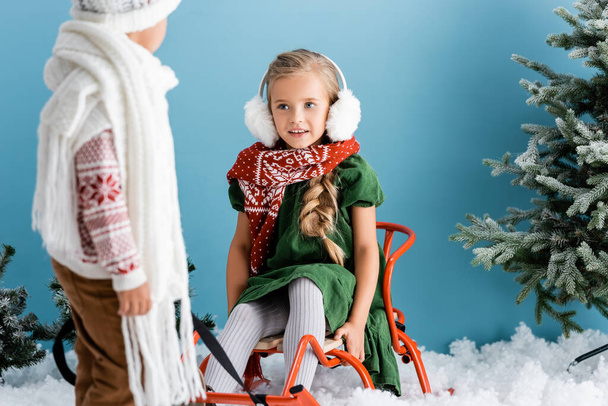 selective focus of girl in winter earmuffs sitting on sleigh near brother on blue - Photo, Image