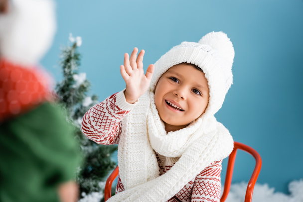 selective focus of boy in knitted scarf and hat waving hand on blue - Photo, image