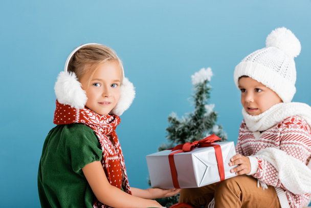selective focus of girl in winter earmuffs near boy in knitted hat holding present on blue - Foto, Bild