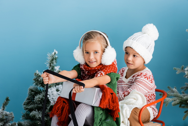 Kinder im Winteroutfit fahren Schlitten mit Geschenk in der Nähe von Kiefern auf blau - Foto, Bild