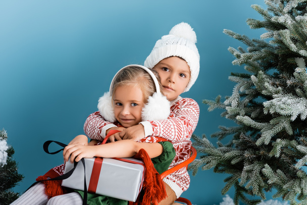 Bruder im Hut umarmt Schwester, während sie in der Nähe von Kiefern mit Geschenk auf blau sitzt - Foto, Bild