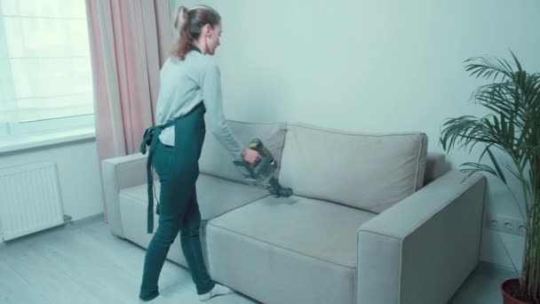 Beautiful young woman vacuums the gray sofa from dust. - Záběry, video