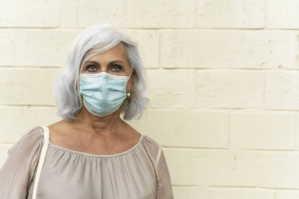 žena v šatech stojí u zdi v ochranné masce. život ženy během koronaviru - Fotografie, Obrázek