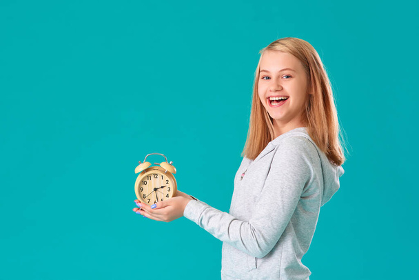 A teenage girl holds an alarm clock, standing on an isolated turquoise background, very happy and excited, the expression of a winner , she celebrates the victory by shouting with a big smile. - Photo, Image