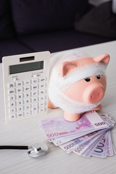 Selective focus of piggy bank with bandage on money near calculator  - Photo, Image