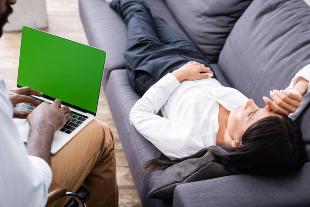 selektivní zaměření africké americké psycholog pomocí notebooku se zelenou obrazovkou v blízkosti ženy ležící na pohovce - Fotografie, Obrázek