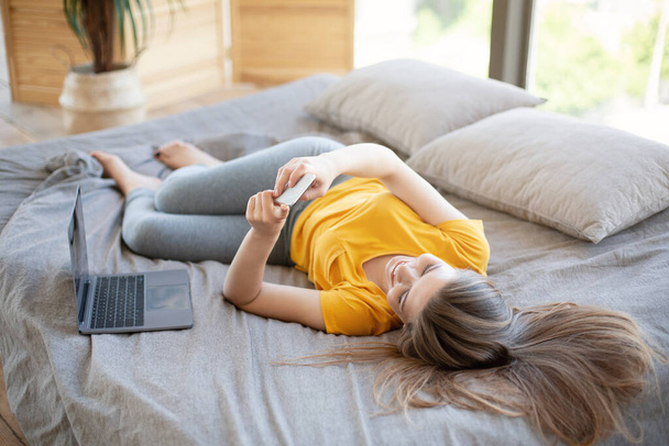Menina milenar feliz com smartphone e computador portátil comunicando on-line na cama dentro de casa - Foto, Imagem