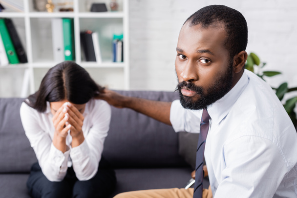 ernstige afrikaanse amerikaanse psycholoog kijken naar camera terwijl kalmerende huilende vrouw verhullen gezicht met handen - Foto, afbeelding