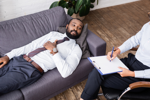 високий кут зору афроамериканця в офіційному одязі, лежить на дивані біля психолога, який пише на кишені
 - Фото, зображення