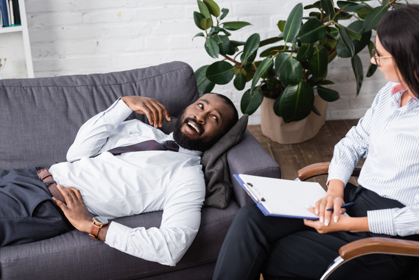 Χαρούμενος Αφροαμερικάνος που μιλάει με ψυχολόγο ενώ ξαπλώνει στον καναπέ. - Φωτογραφία, εικόνα