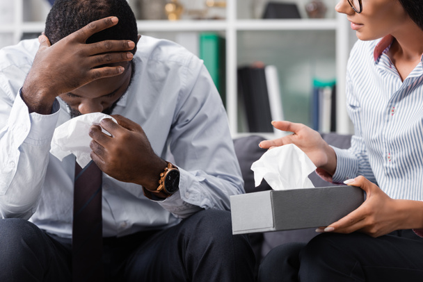 psicólogo sosteniendo servilletas de papel cerca estresado paciente afroamericano llorando y tocando la cabeza en el hospital - Foto, imagen
