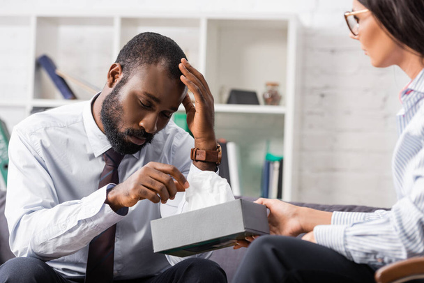 селективный фокус разочарованного африканского американца касающегося головы во время приема бумажной салфетки от психолога - Фото, изображение