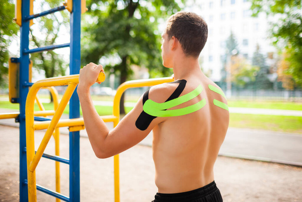 Unrecognizable young caucasian professional bodybuilder with black and green elastic tapes on shoulders posing at sports ground. Man with naked back holding iron bars outdoors. - Photo, Image