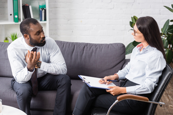selective focus of worried african american man gesturing while talking to brunette psychologist holding clipboard - Fotó, kép