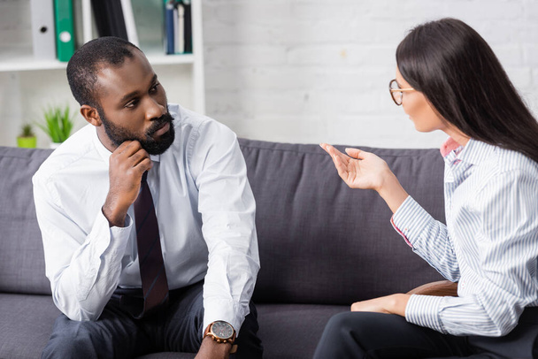brunette psychologist gesturing while talking to concentrated african american man holding hand near face - Photo, Image