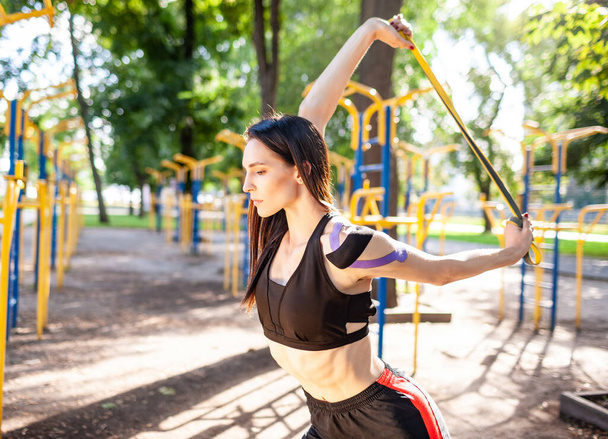Brunette muscular woman posing with fitness resistance band in park, sports ground on background. Back view of young female with elastic taping on body training outdoors. Rehabilitation concept. - Foto, Imagem