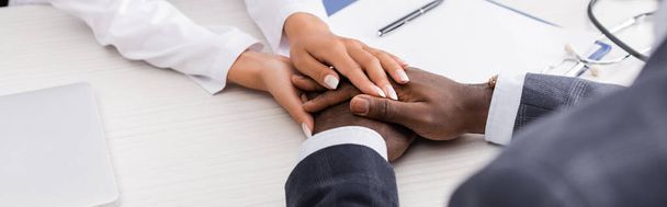 vista parcial del médico tocando las manos del hombre afroamericano cerca del portapapeles y la pluma en el escritorio, imagen horizontal - Foto, imagen