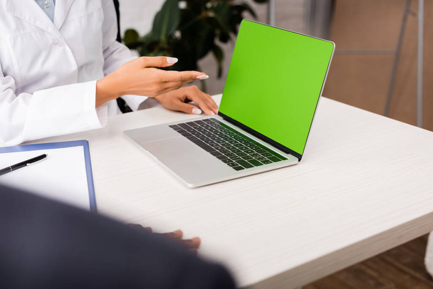 oříznutý pohled lékaře ukazujícího rukou na notebook se zelenou obrazovkou v blízkosti pacienta na pracovišti - Fotografie, Obrázek