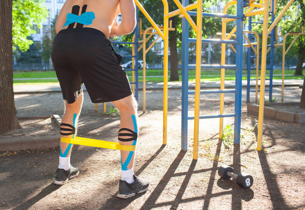 Muscular man training fitness resistance band on legs at sports ground. Back view of young unrecognizable bodybuilder with elastic kinesiology taping on body training outdoors. Rehabilitation concept. - Фото, зображення