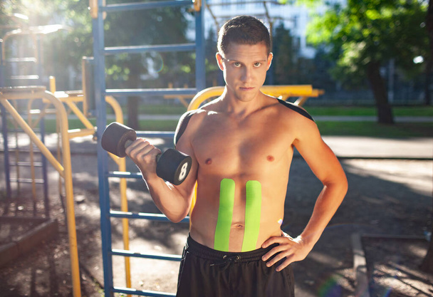 Portrait of handsome young caucasian professional bodybuilder with black and green elastic tapes on body carrying dumbbell at sports ground. Man with muscular naked torso looking at camera. - Foto, immagini