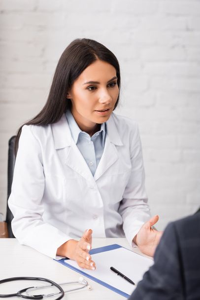 selective focus of doctor talking to patient while sitting at workplace near stethoscope and  clipboard - Fotografie, Obrázek