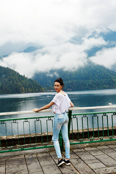 Dziewczyna w białym ubraniu na tle górskiego jeziora z błękitną wodą i górami z chmurami. Kontekst. - Zdjęcie, obraz