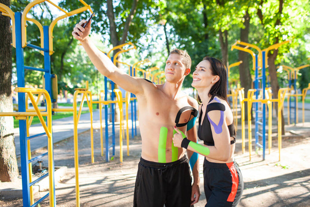 Professional caucasian athletes with kinesiology elastic taping on bodies, handsome man and brunette woman, posing at sports ground, having rest and taking selfie using smartphone. - Photo, image